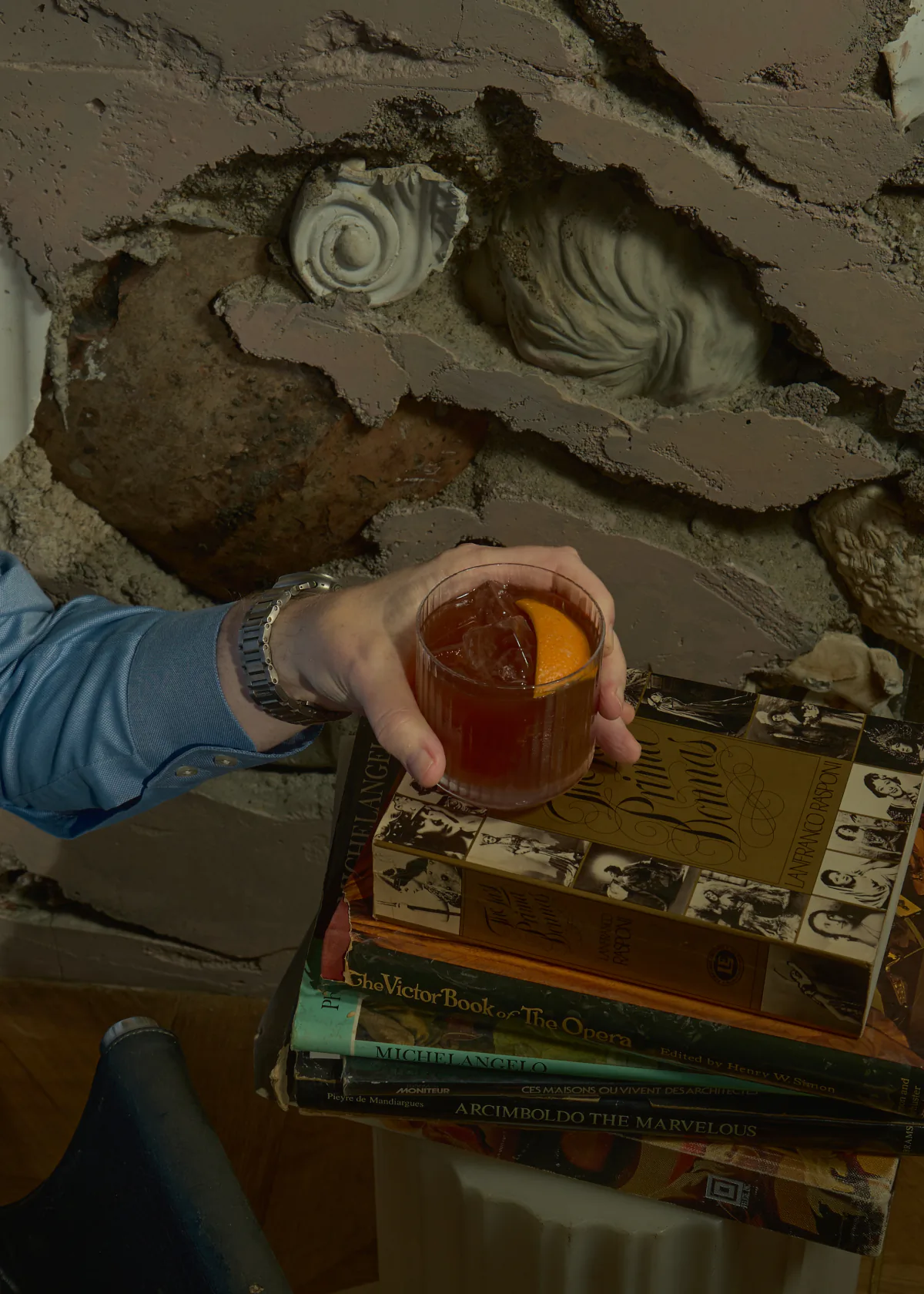 A hand rests a negroni on a stack of art books in front of a wall of cast concrete with carved stone fragments set into it