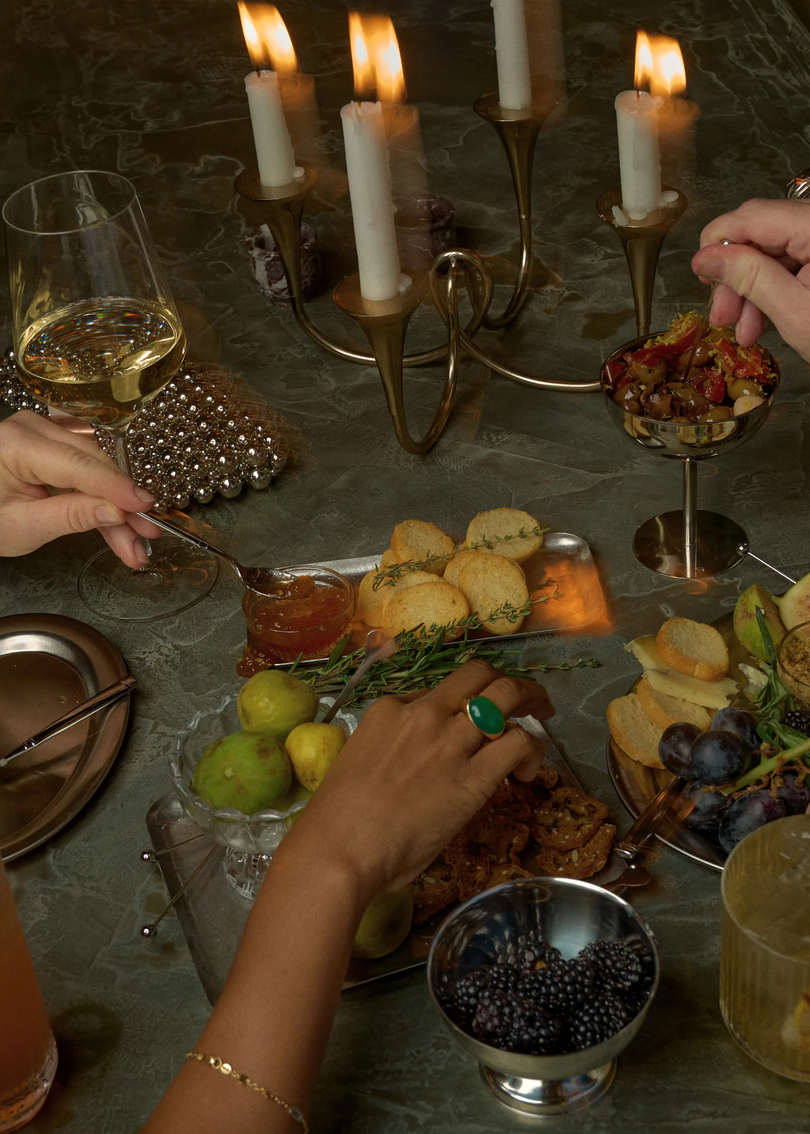 Hands reach across a candlelit table set with olives, crackers, blackberries, figs and a glass of white wine
