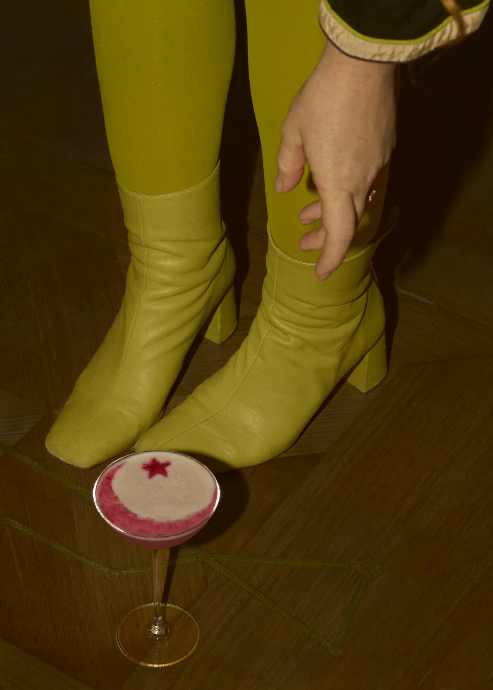 Chartreuse boots and tights on a parquet floor, with a pink cocktail set down beside them on the floor