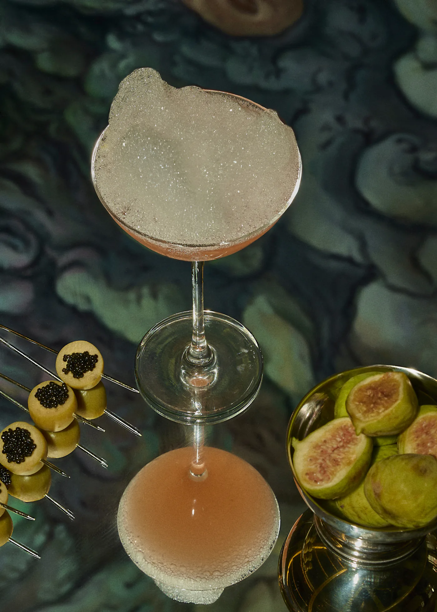 A pink sour with a thick white foam in a coupe, seen from above on a dark marbled table, beside caviar canapes and a silver bowl of split figs