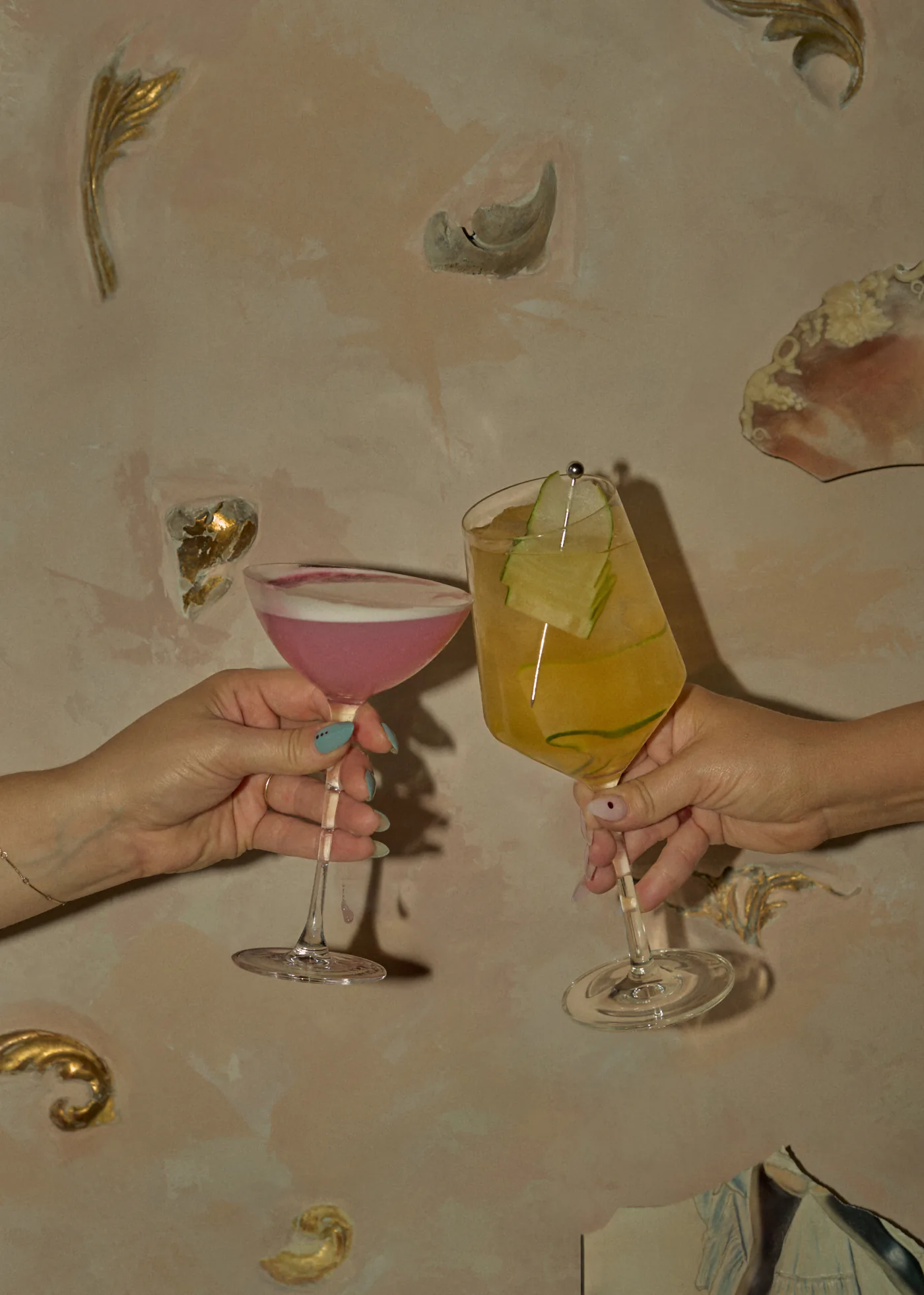 Two hands touch a pink coupe and an amber cocktail together against a pale plaster wall worked with gold leaf
