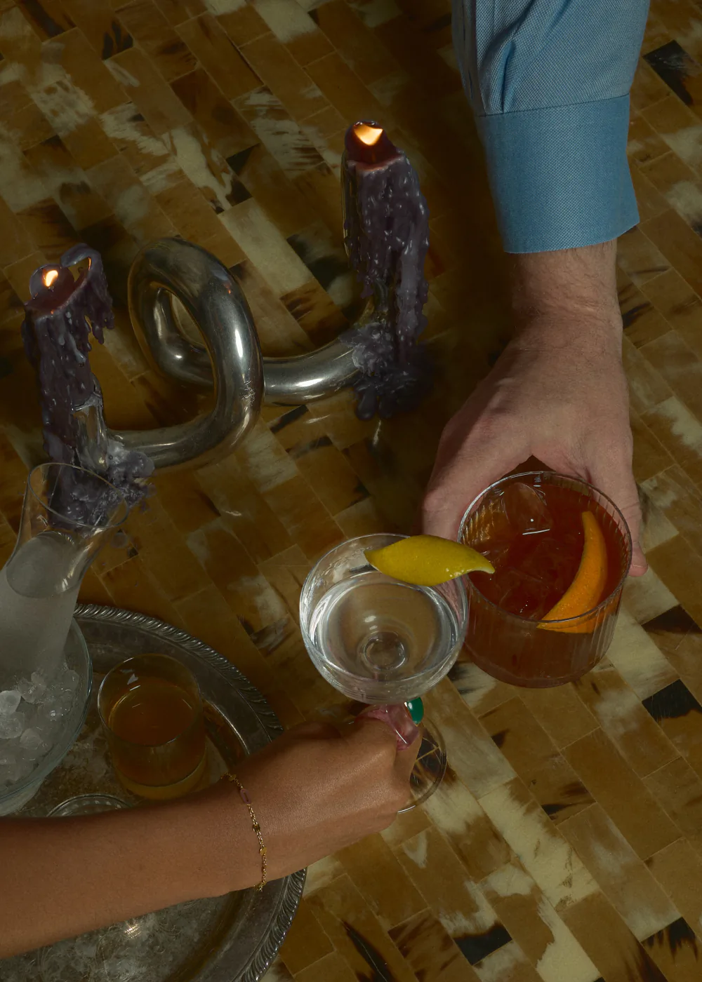 A hand in a blue cuff sets down an amber cocktail beside a lit candle and a coupe on a tiled table