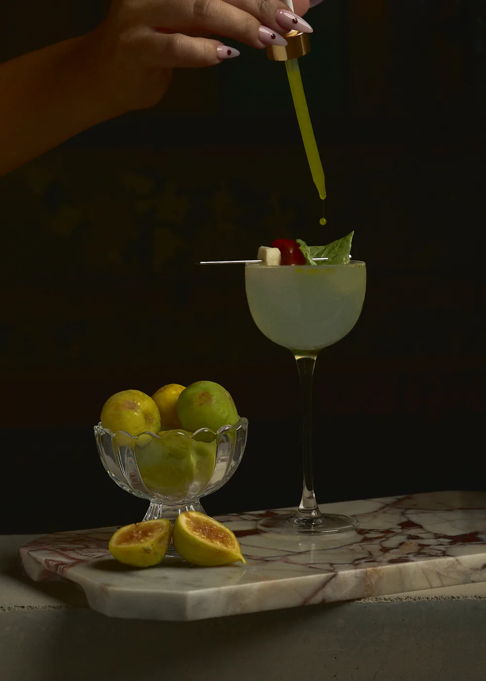 A hand lowers a skewer of basil, tomato and cheese onto a pale green cocktail in a coupe, on a slab of marble with a bowl of figs
